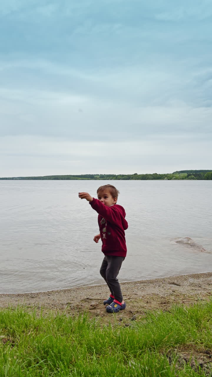 Happy Caucasian toddler stands at the river bank. Kid throws the pebbles into water and picks some more from the shore. Vertical video.