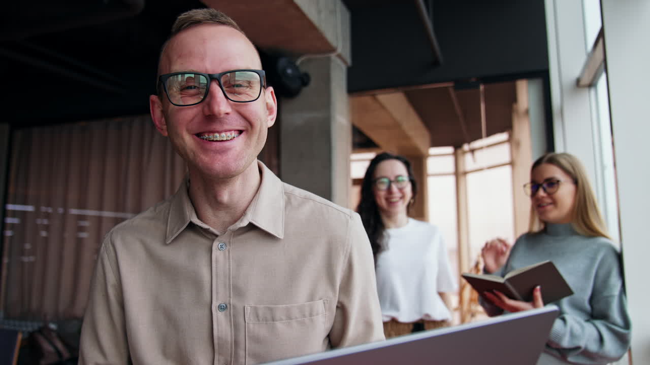 Laughing Caucasian man holding a laptop. Female colleagues discuss work issues at backdrop.