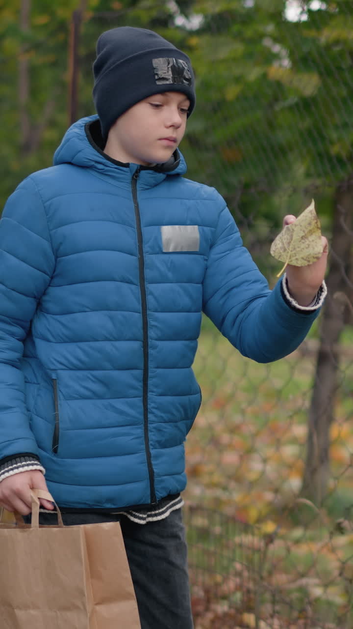 파란색 재과 베니를 입은 러시아 소년은 땅에서 노란 잎을 뽑기 위해 몸을 숙이고 활기찬 가을 잎자루와 낙 잎 사이에서 종이 봉투에 넣습니다.