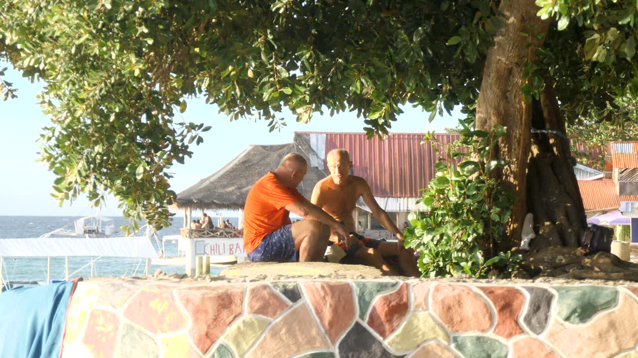 Elderly Men Having Conversation Under The Shade Of A Tree Near Shoreline Of Moalboal In Cebu, Philippines. Static Shot