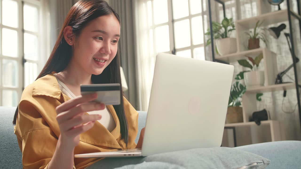 Happy Asian Woman Sitting On Couch In Living Room Holding Credit Card And Using Laptop Computer Shopping Online