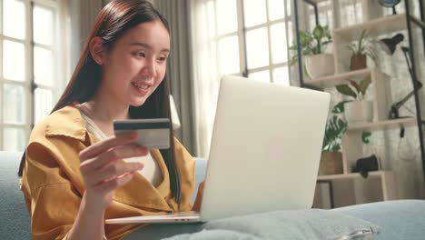 happy asian woman sitting on couch in living room holding credit card and using laptop computer shopping online