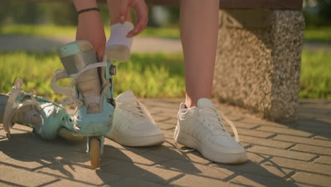 partial view of individual reaching for roller skate, preparing to put it on after removing right leg from sneaker, white sneakers on feet and grass in background with sunlight casting soft