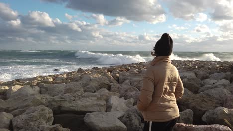 a girl stands on the rocks watching the large waves of a wild sea on a windy day in europe