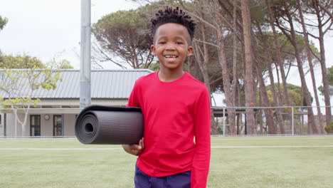 african american boy carrying yoga mat on school field, looking excited outdoors