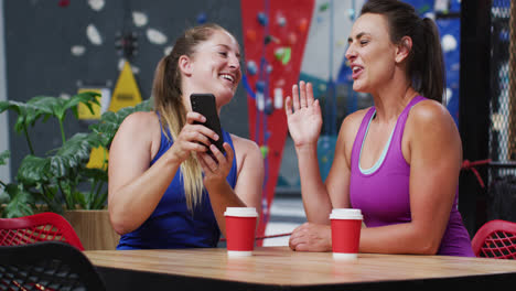 Dos-Mujeres-Caucásicas-Felices-Riendo-Y-Mirando-El-Teléfono-Inteligente-En-La-Cafetería-En-El-Muro-De-Escalada-Interior