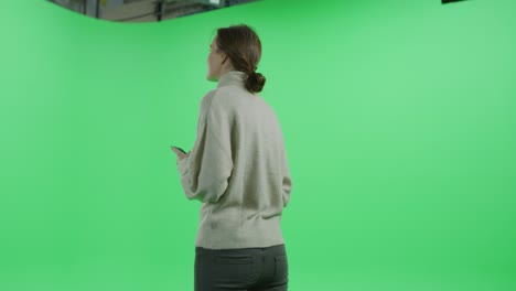 green screen chroma key studio: beautiful young girl using smartphone, looks around in wonder, browsing through internet and social media. 360 degree tracking arc shot