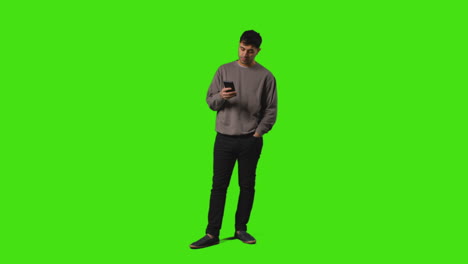 joven enviando o leyendo un mensaje de texto o navegando en línea en un teléfono móvil de pie contra la pantalla verde