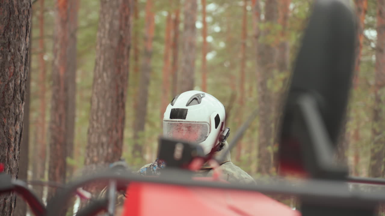 Un pilota caucasico ispeziona un quad rosso in una pineta, indossando un casco bianco e una giacca mimetica, primo piano del viso concentrato e della visiera, il pilota controlla i comandi e gli indicatori accanto ad alberi spolverati di neve, luce fredda e silenzio.
