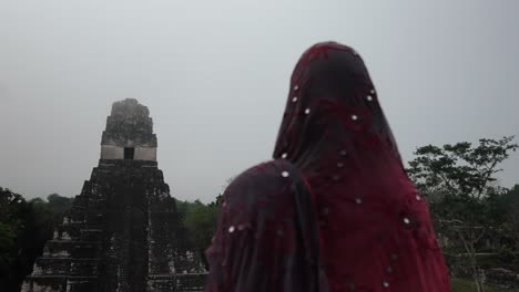 a woman in a decorated shawl gazes at temple i in tikal as the misty jungle creates a mystical atmosphere.