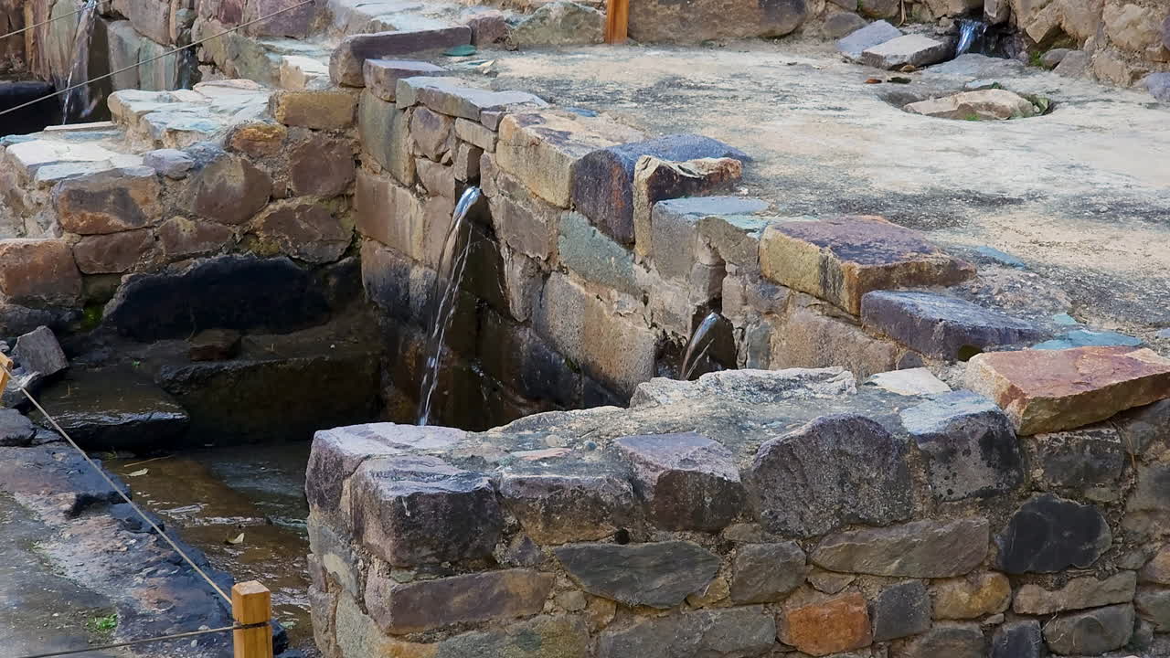 A cinematic pan shot captures the ancient Inca water fountains and intricate stone channels of the Ollantaytambo Archaeological Park, showcasing their historic and cultural significance