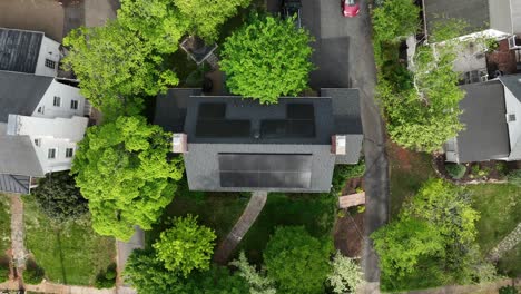modern house with solar panels on roof between green trees in spring. american suburb neighborhood producing green energy. aerial top down rising shot.