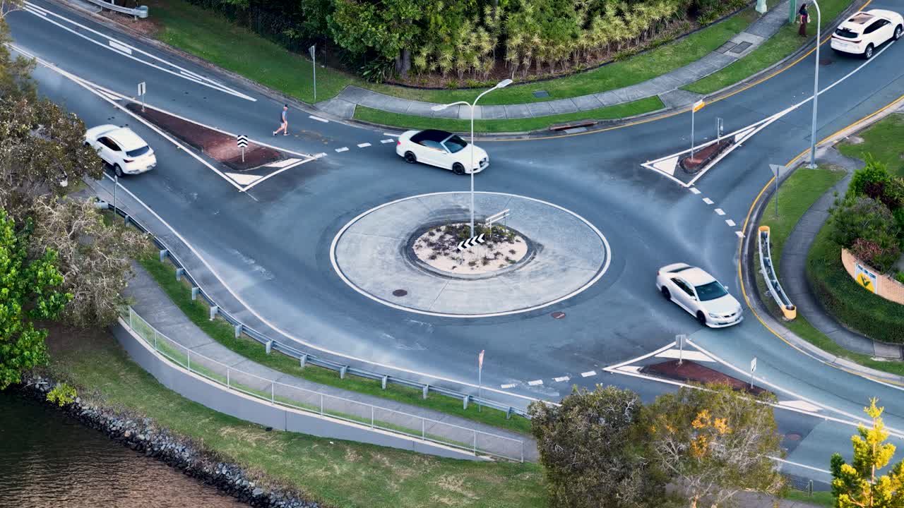 Drone footage captures cars navigating a roundabout surrounded by greenery in Gold Coast, Australia, under natural daylight
