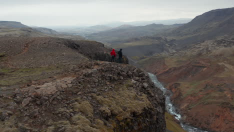 Drone-view-people-hikers-standing-top-cliff-looking-stunning-panorama-in-Iceland.-Aerial-view-two-tourist-contemplating-amazing-Landmannalaugar-valley-with-fossa-river-flowing-mossy-highlands