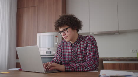 un hombre rizado con una mirada seria trabaja en una computadora portátil sentado en una cocina moderna. joven estudiante independiente usando computadora portátil estudiando en línea trabajando desde casa en internet