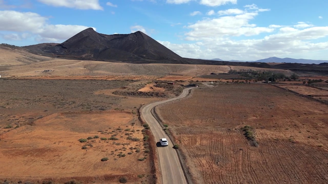 Auto bianca che percorre una strada nel deserto a Fuerteventura con un vulcano sullo sfondo, che evoca un concetto di viaggio e road trip