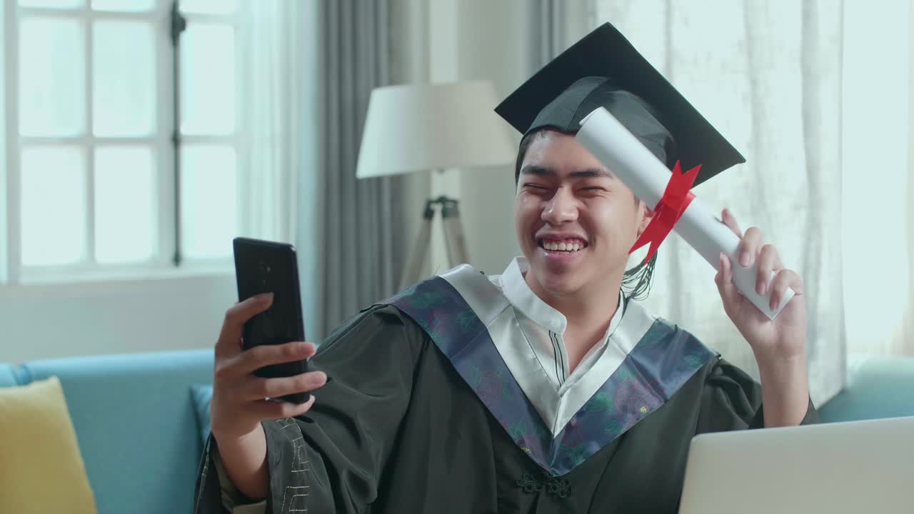 Excited Asian Man Showing Off A University Certificate To The Family During An Online Video Call By Mobile Phone. Male Graduate Wearing A Graduation Gown And Cap Sitting On The Living Room