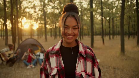 Portrait-of-a-happy-girl-with-black-skin-in-a-checkered-shirt-who-looks-at-the-camera-and-poses-against-the-background-of-other-participants-in-a-hike-and-tent-in-a-Sunny-summer-green-forest