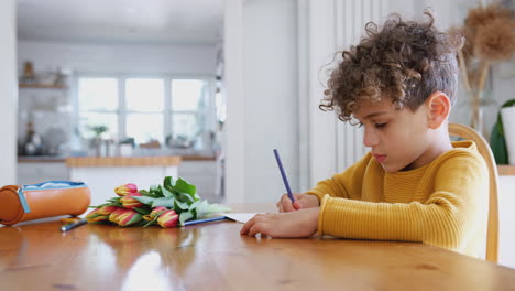 young boy at home with bunch of flowers writing in mothers day card