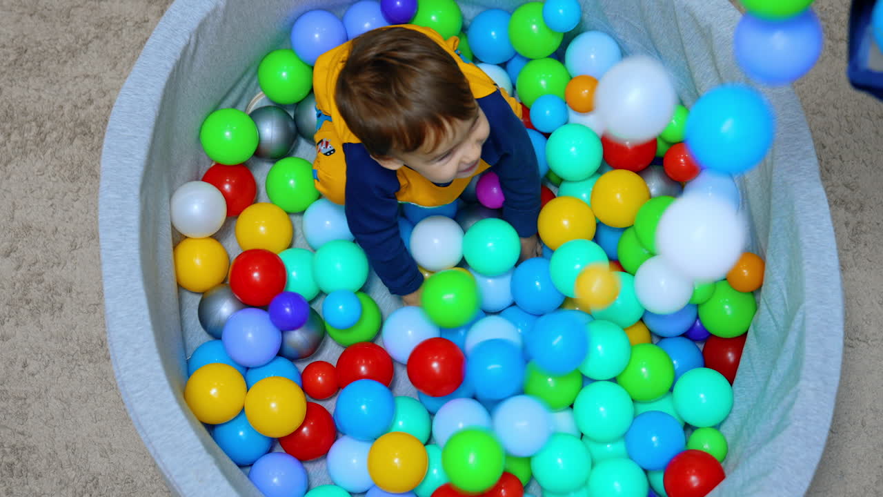 Little boy sits in the dry pool. Somebody pours the plastic balls on the kid and the child is smiling. Top view.