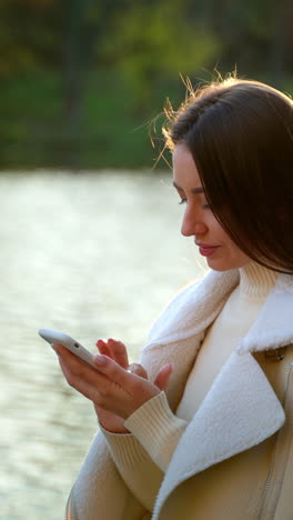 brunette lady focused on her phone standing at the bridge. pensive woman typing a message on smartphone in autumn season. blurred backdrop.