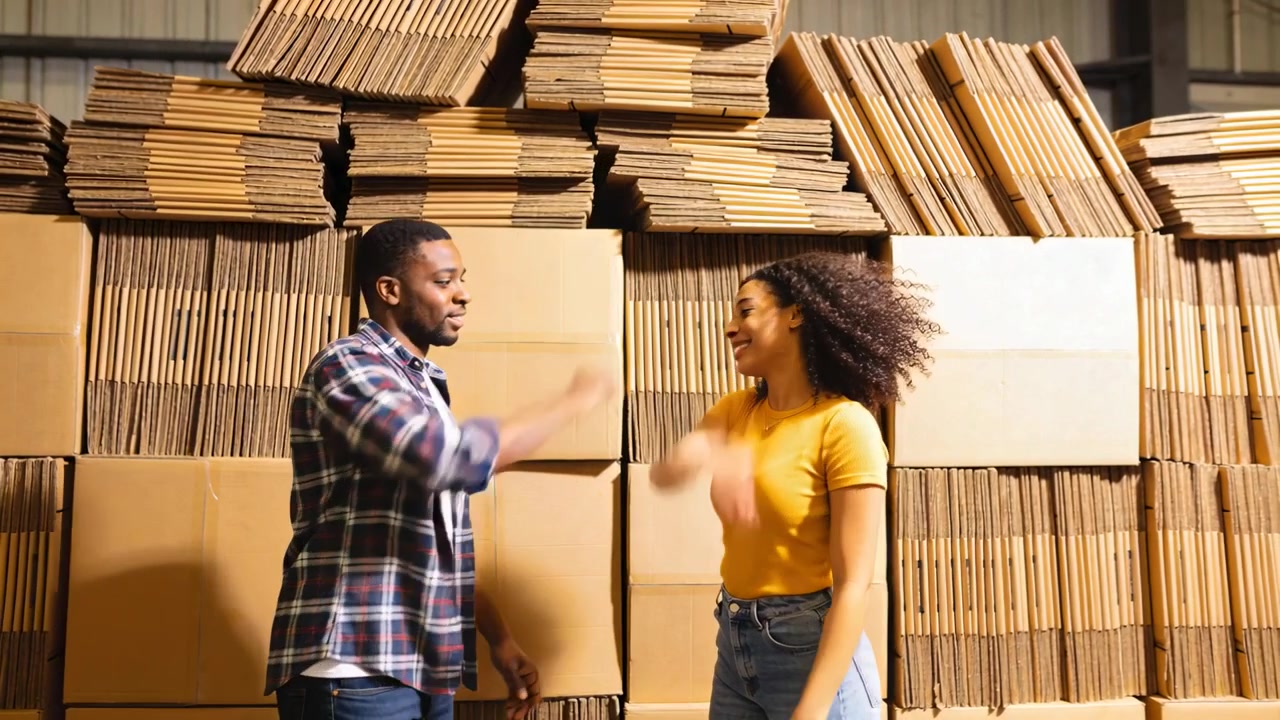 High-fiving coworkers in plaid, yellow tee after packing at warehouse, with cardboard stacks
