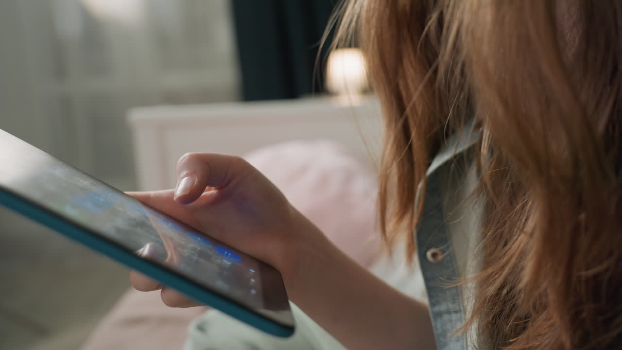 Primo piano di mani che tengono un tablet, giovane studente che scorre app su un tablet blu mentre è seduto sul letto in camicia di jeans, luce soffusa della lampada e cuscino pastello sullo sfondo, atmosfera serale rilassata, tocco del polpastrello.