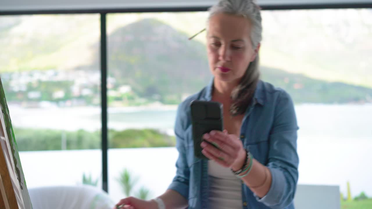 Donna in jeans prende un pennello, controlla il telefono per riferimento e dipinge su un cavalletto vicino a una finestra con vista sul mare.