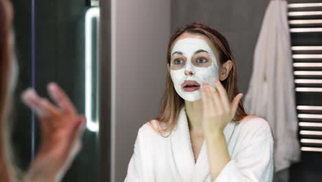 Blonde-woman-in-bathrobe-putting-white-mask-for-moisturizing-using-fingers