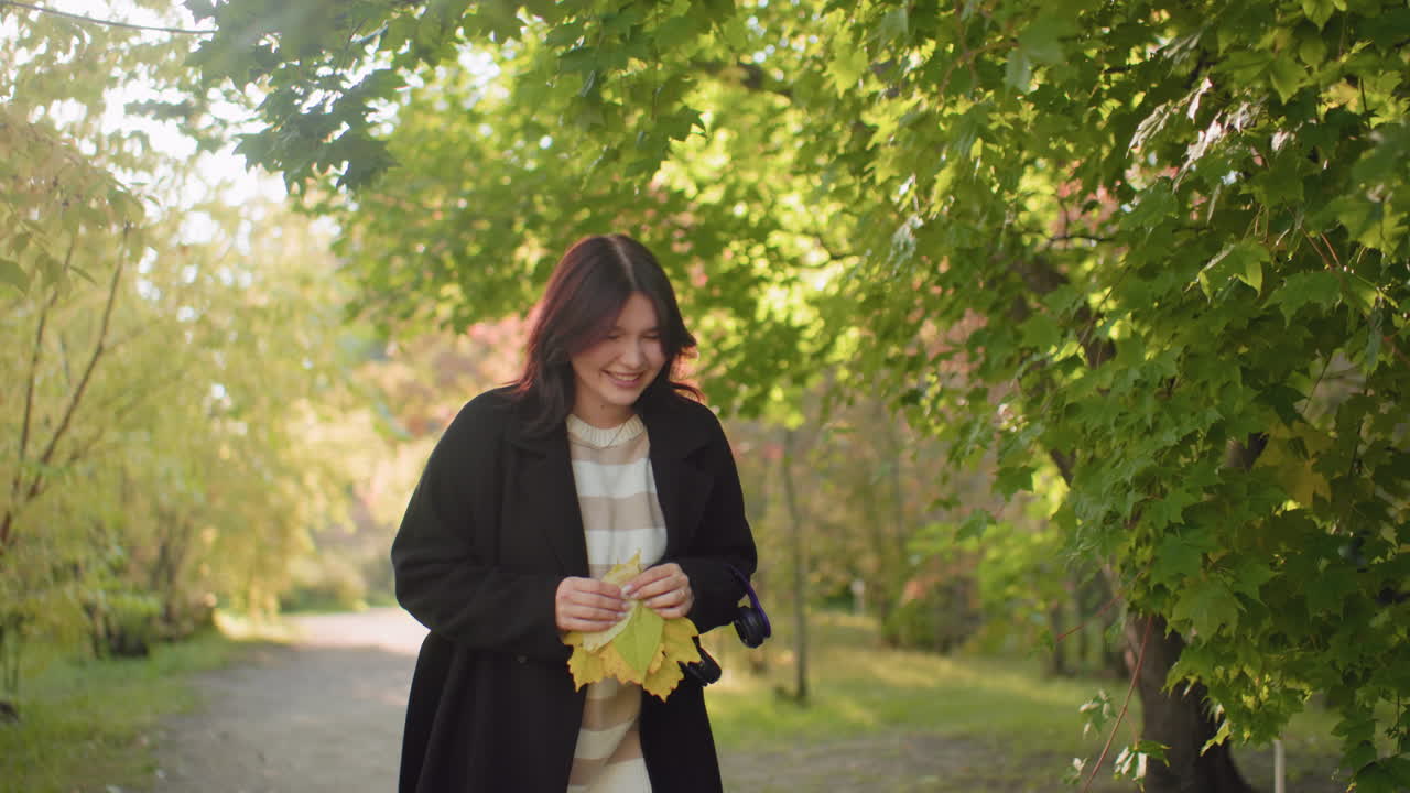 Beautiful young girl standing with bunch of autumn leaves in hands, moving body with excitement, smiling under warm sunlight in forest pathway surrounded by green trees and seasonal foliage