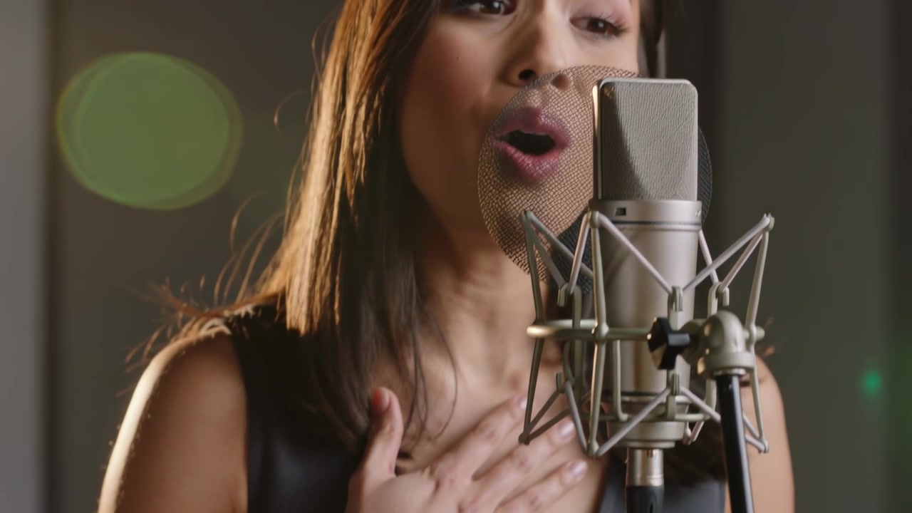 Cantante asiatica che prende fiato, indossa un top senza maniche, preme una mano sul petto e si china verso il microfono mentre registra una canzone.