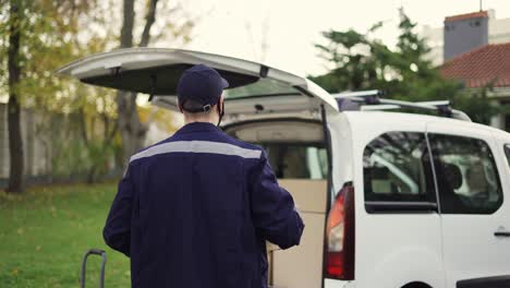 handsome courier putting cardboard box package in delivery van and enroll the parcel. courier in mask, cap and gloves on the way