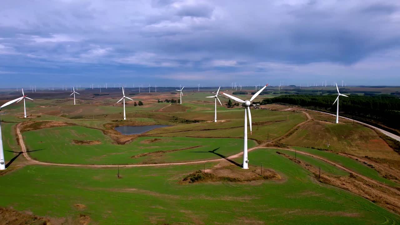Rotating Wind Turbines In Hilly Landscape Against Blue Cloudscape Sky