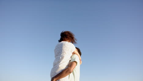 biracial couple embraces under a clear blue sky, woman's curly hair blowing in the wind