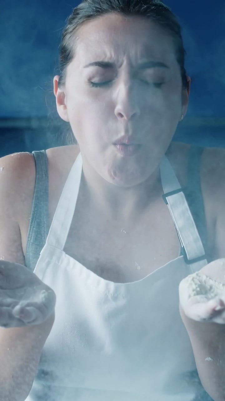 Vertical video: Cupping handfuls, woman in white apron blowing flour in kitchen, dusting for baking