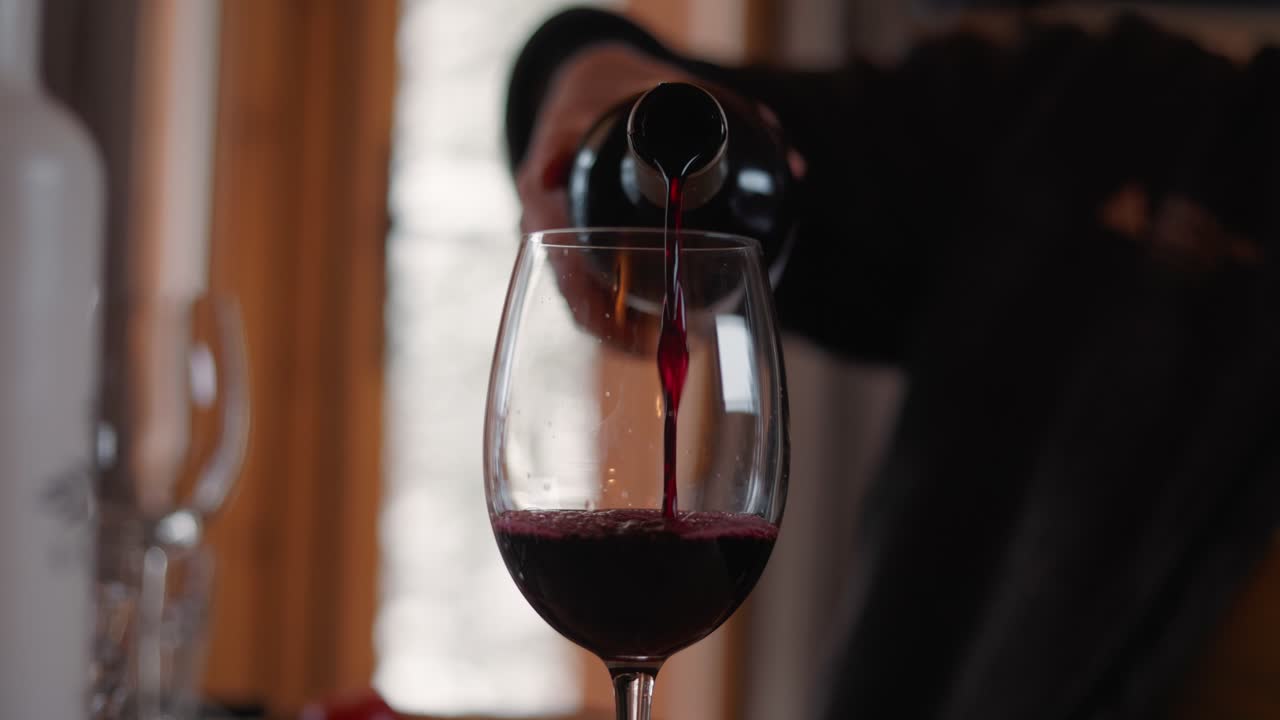 Red wine being poured into a glass. Close-up shot indoors, showing only the hand and arm of the person serving.