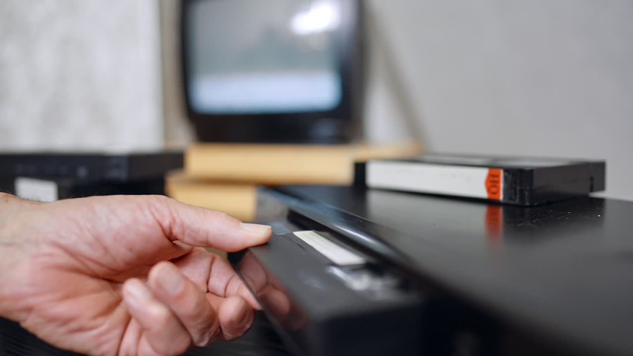 VHS player, several tapes and shimmering screen. A cassette appears from the player and hand puts it on the pile