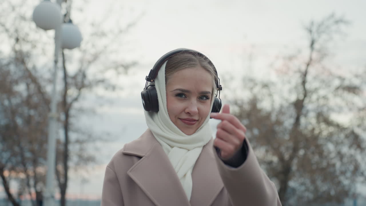 Donna in cappotto che fa un gesto a cuore alla telecamera, con cuffie wireless e sciarpa di lana, sguardo amichevole. Vibrazioni da influencer dei social media e invito alla connessione.