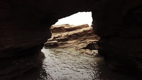 drone aerial zoom out from a small rocky arch way to reveal ocean and beach