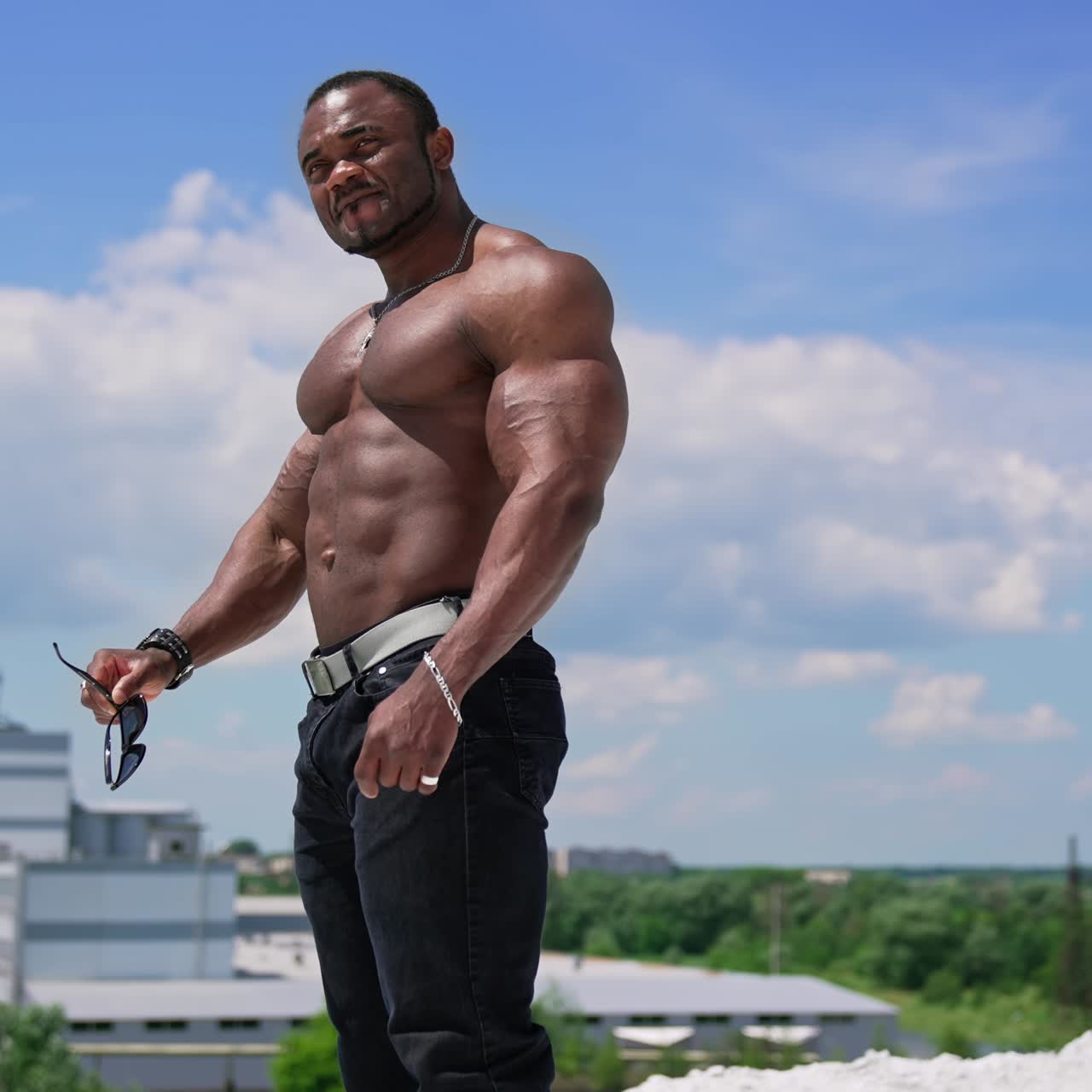 Portrait of handsome african sportsman outdoors. Cheerful shirtless athlete with strong trained muscles standing on the hill against blue sky
