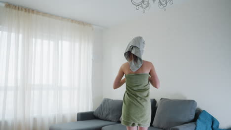back view of lady wrapped in light green towel with hair covered by cloth walking toward couch in bright cozy living room with soft natural light through sheer curtains and stylish chandelier