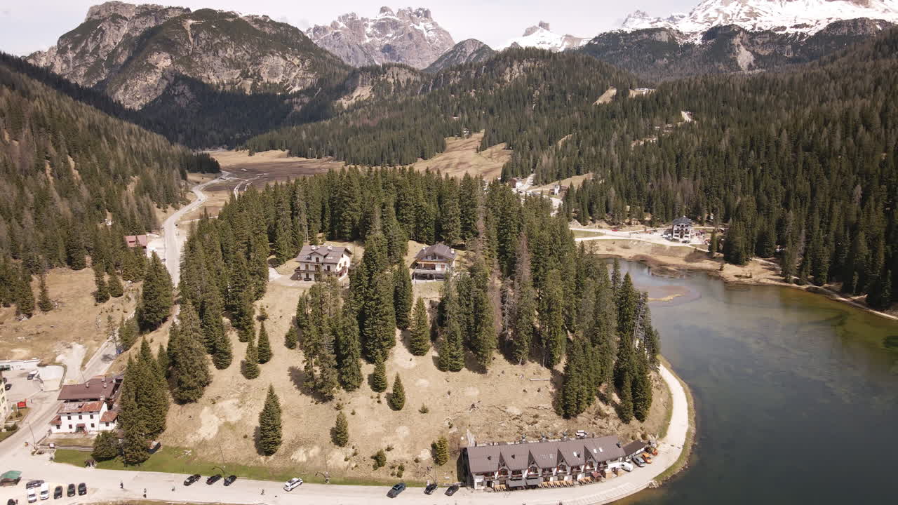 Camera pulls back from secluded chalets nestled among dense evergreens, revealing a serene alpine lake, winding valley road, and towering mountain backdrop