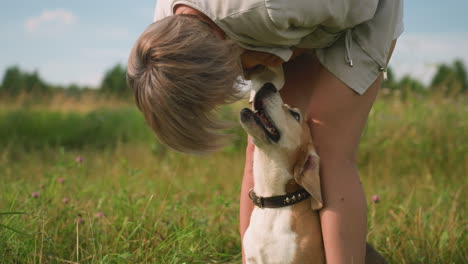 dog owner bending down in grassy field, playfully interacting with her dog while dog looks up lovingly, leash around dog's neck, sunny outdoor setting with lush green grass