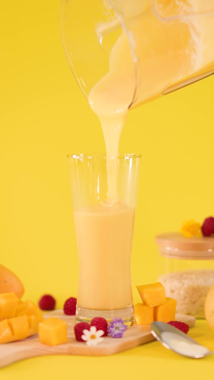 Smoothie al mango al rallentatore versato in un bicchiere alto con una composizione di frutta tropicale e illuminazione brillante da studio.