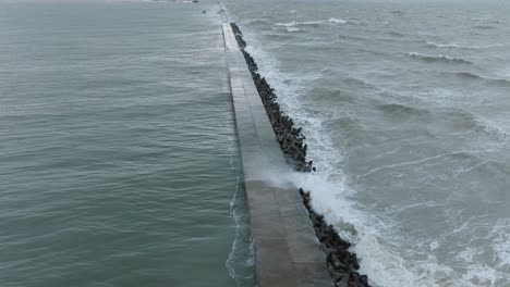 aerial establishing view of port of liepaja concrete pier , autumn storm, big waves splashing, overcast, wide birdseye drone shot moving forward