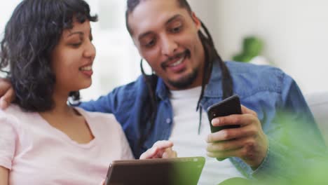 Feliz-Pareja-Birracial-Sentada-En-El-Sofá-Usando-Tableta-Y-Teléfono-Inteligente,-Besándose-En-La-Sala-De-Estar