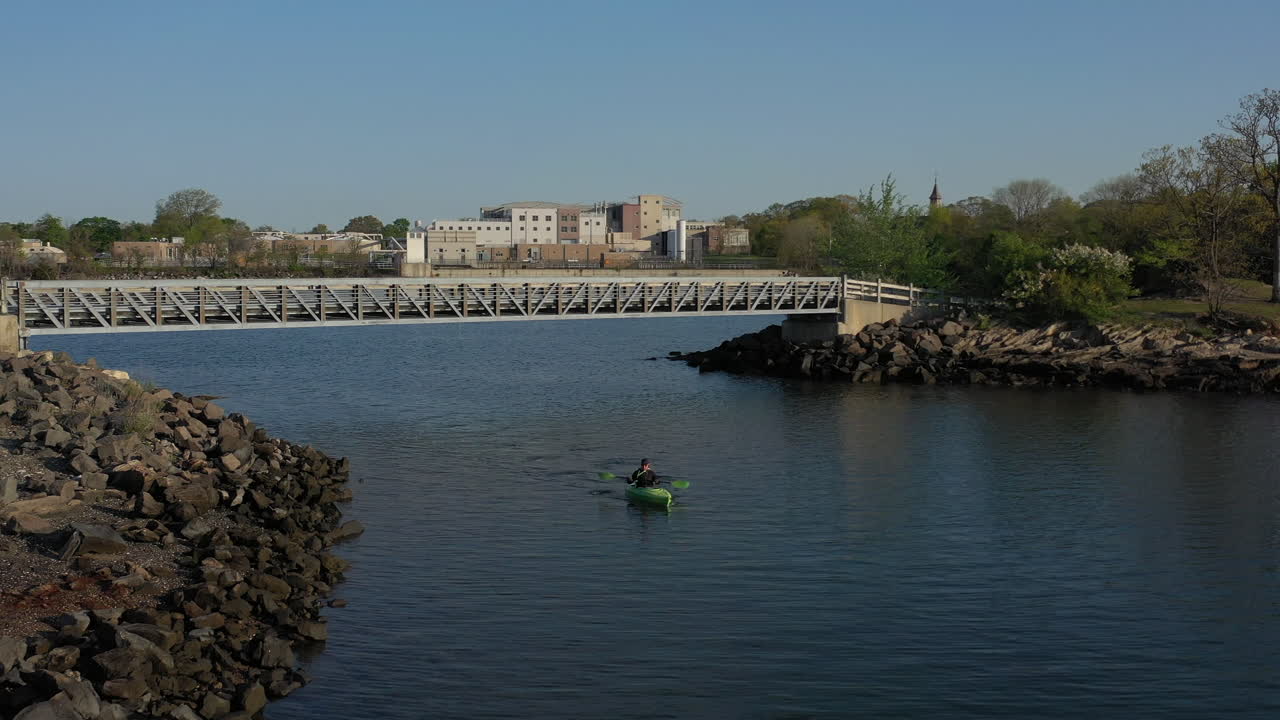 화창한 날 공원 옆 잔잔한 물에서 카약을 젓는 남자의 낮은 각도 드론 샷