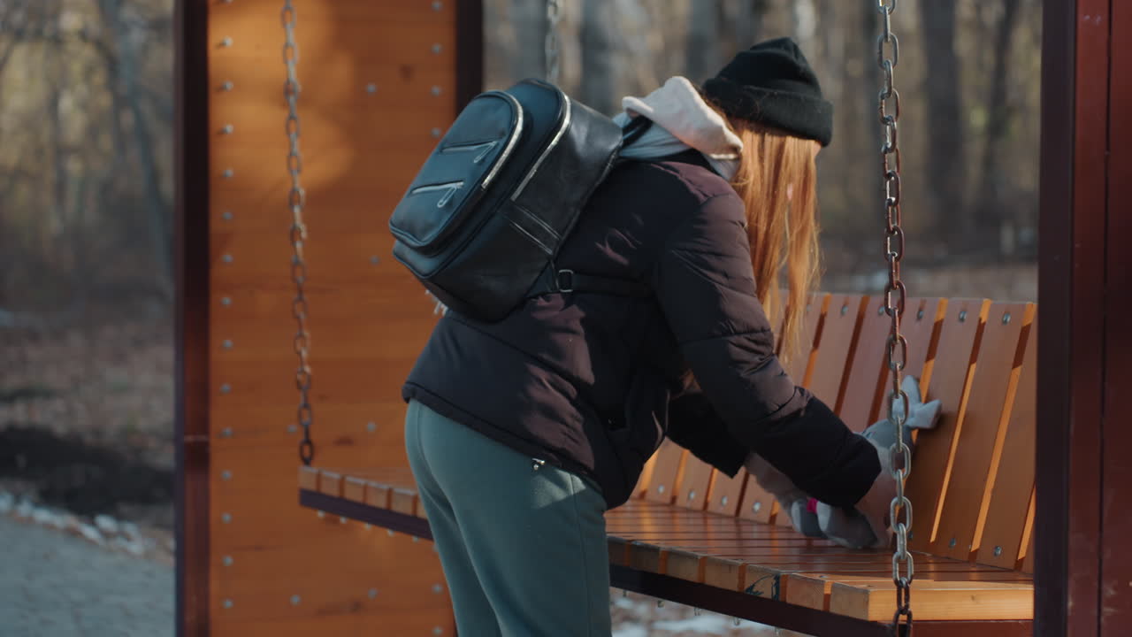 White woman snatcher grabs plush rabbit on wooden swing bench, winter park setting with trees and light snow backpack, beanie and gloves visible as she reaches, conceals toy, and moves furtively