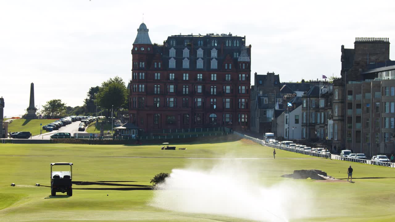 Sprinkler system sprays water on golf course green, morning sunlight, static wide shot, urban backdrop