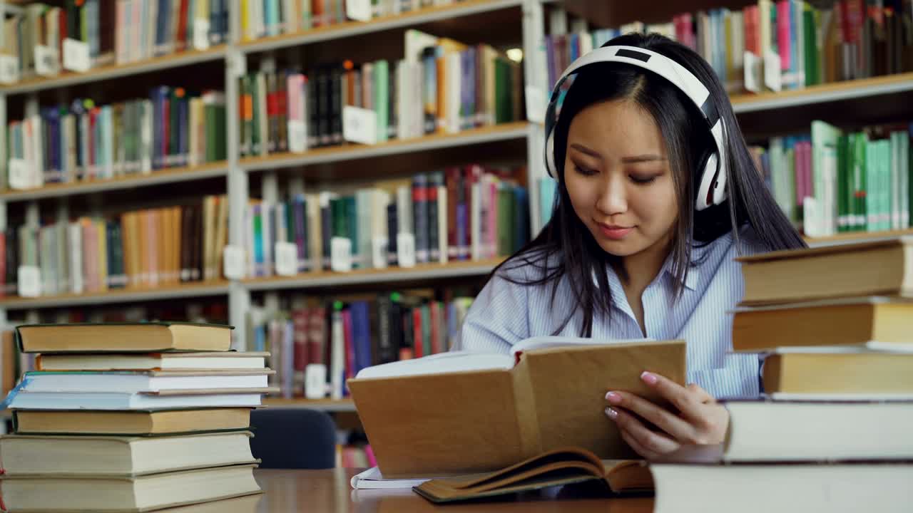 Pretty positive asian teenage student with headphones on head listening to music is sitting at table in big library holding book and writing down summary